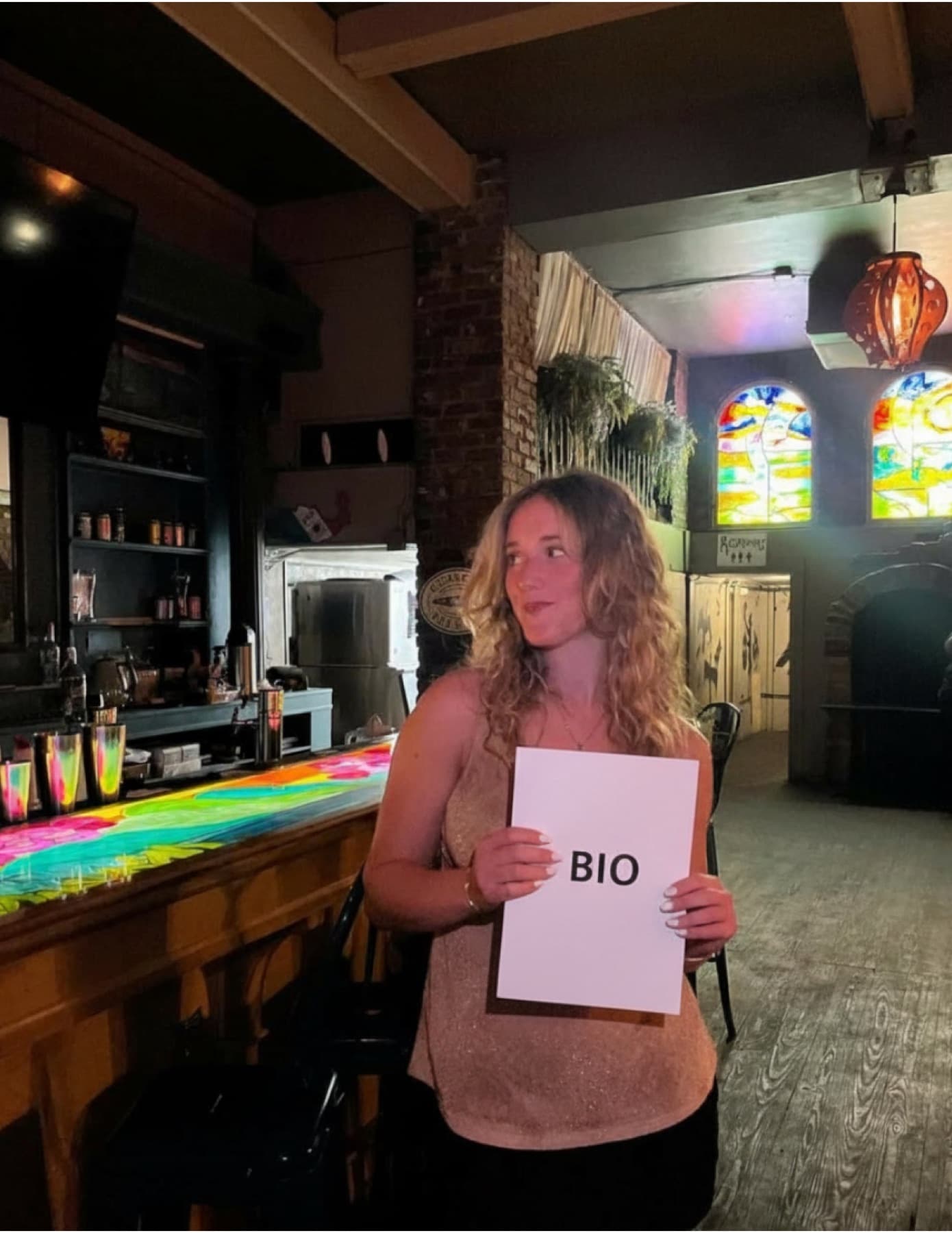 Dahlia Harris standing at a bar with a rainbow-painted counter, holding a hand-lettered sign that reads BIO, stained-glass windows glowing behind her.