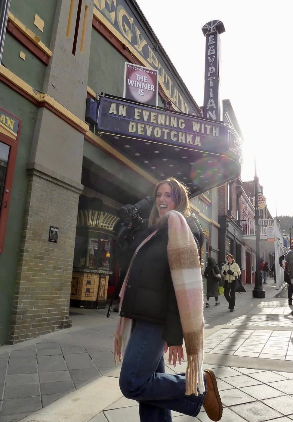 Dahlia Harris laughing mid-step on the sidewalk outside the Egyptian Theatre in Park City, Utah, a camera slung over her shoulder and a striped scarf around her neck.