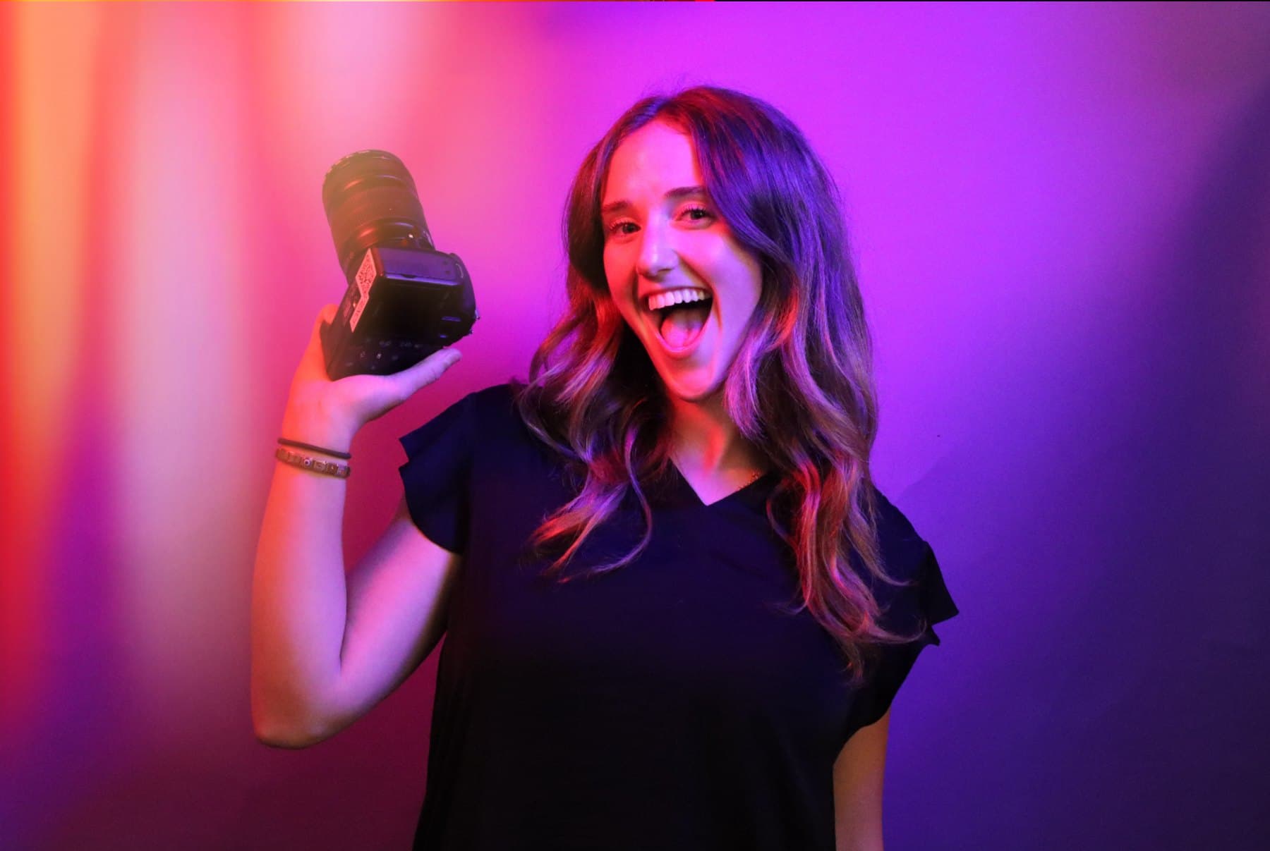 Dahlia Harris laughing and holding up a camera against a wall washed in orange, pink and purple light.